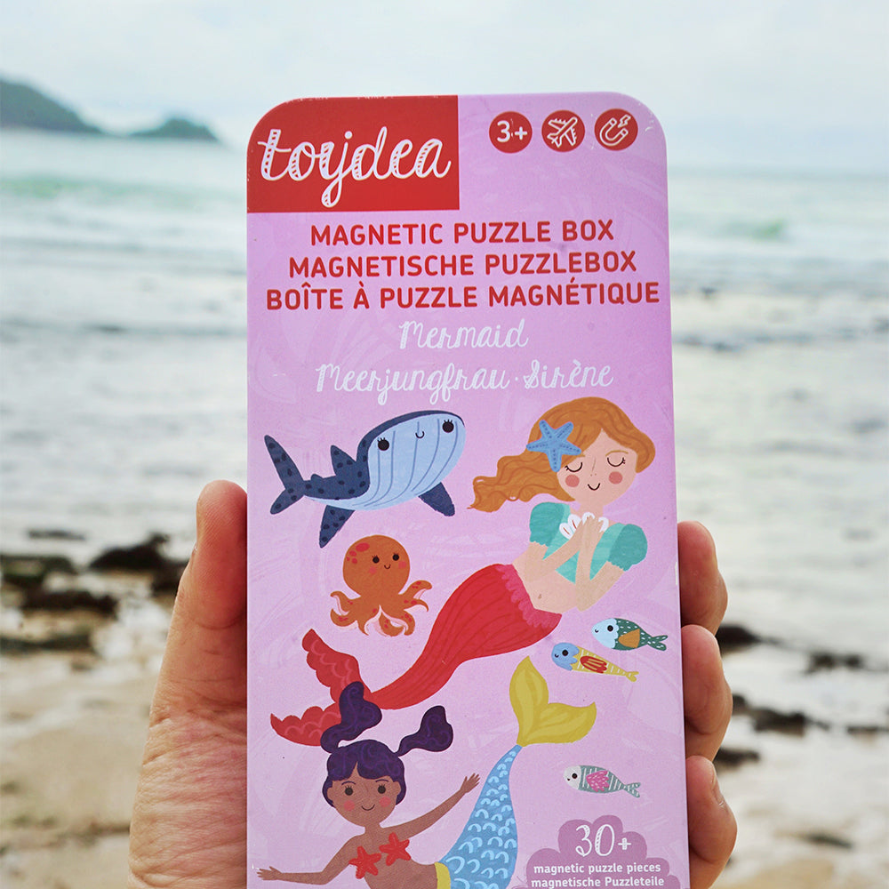 Eine Hand hält die Toydea Magnetspiel-Box 'Meerjungfrau' vor einem verschwommenen Strand im Hintergrund.